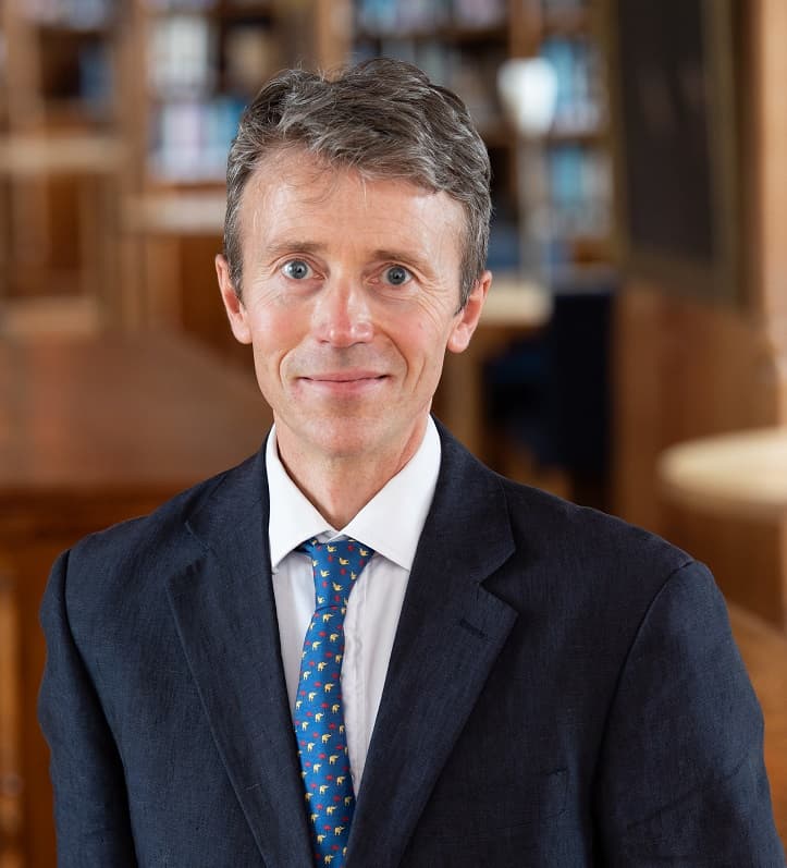 A man in a suit and colorful tie stands in a library, smiling.