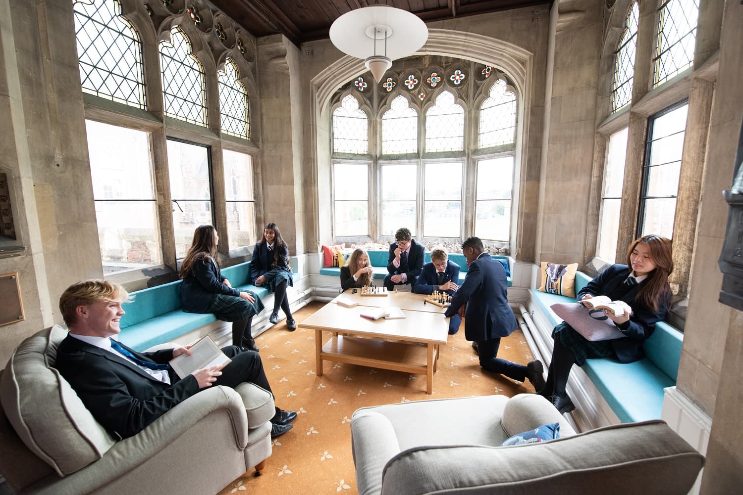 A group of people in formal attire sit and interact in a sunlit, ornate room with large windows and cushioned seating.