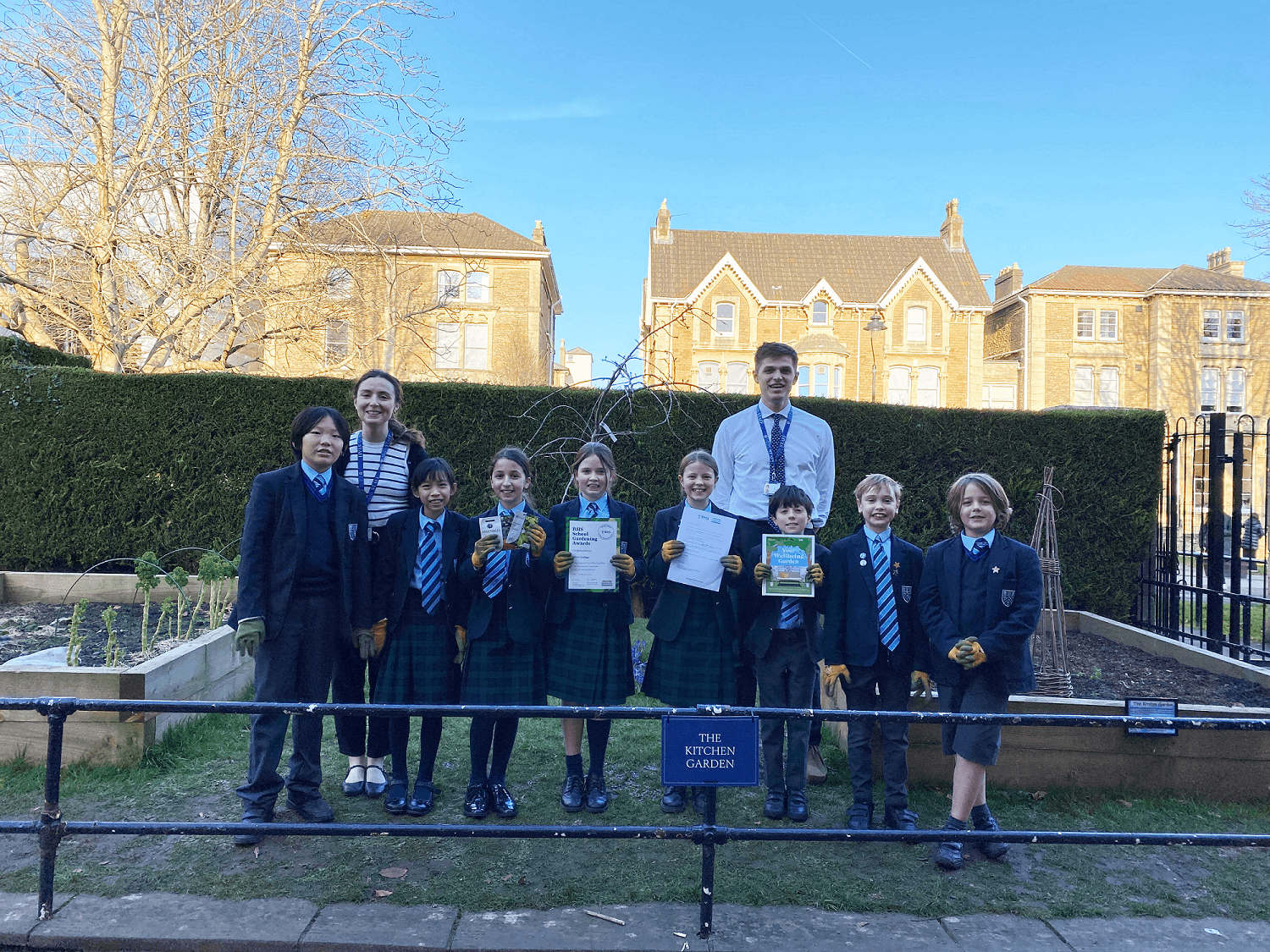 A group of children and two adults stand smiling in a garden, holding certificates. They are dressed in school uniforms.