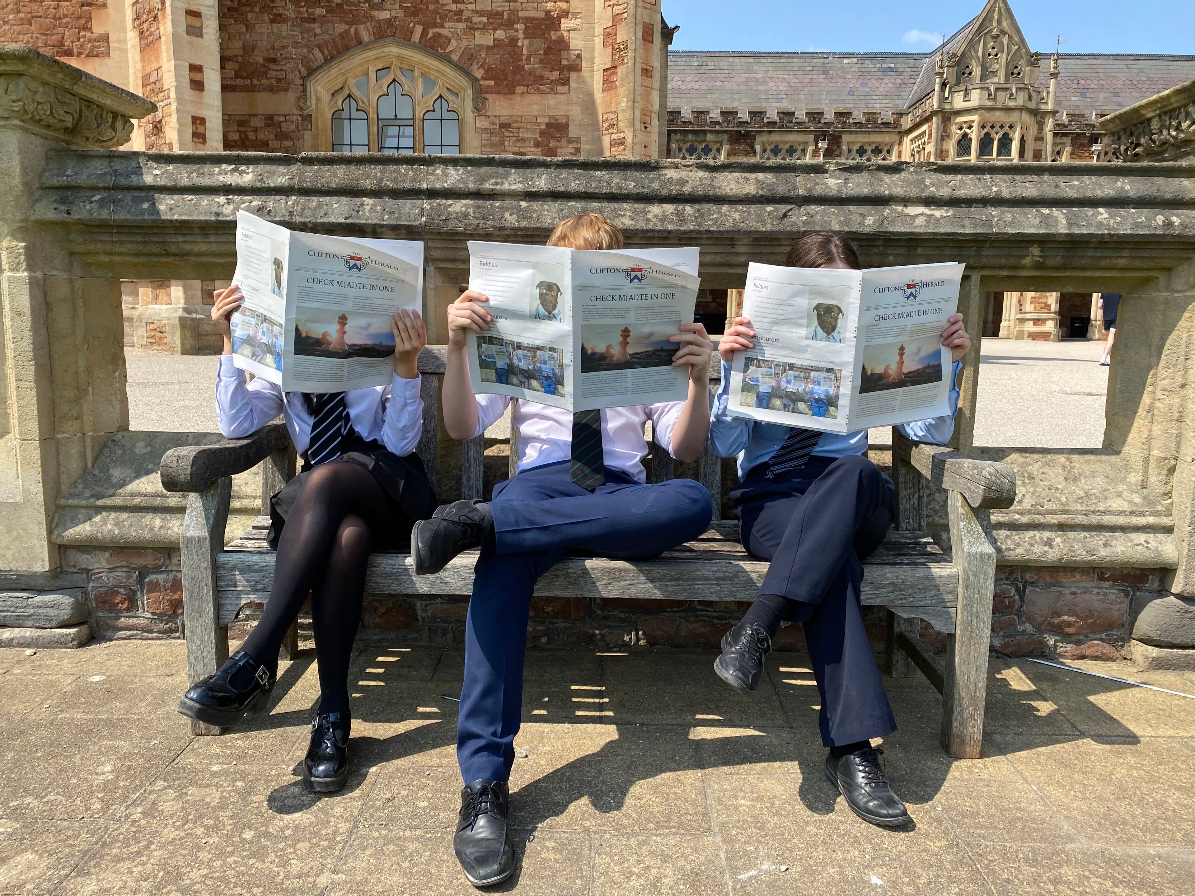 Three people in school uniforms sit on a bench, each reading a newspaper, with a historic building in the background.