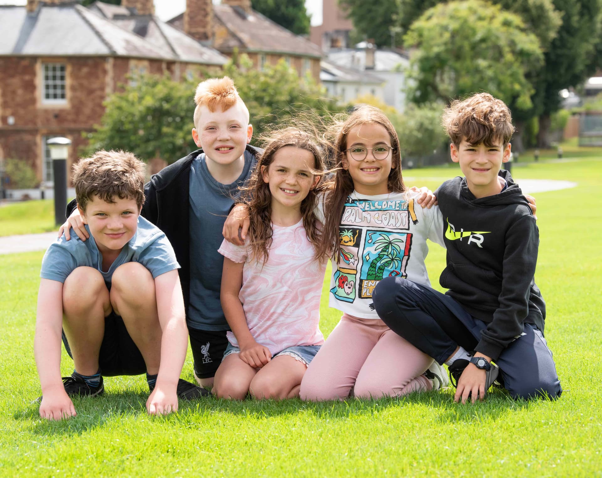 Five children sitting on grass, smiling at the camera. They are outdoors with trees and buildings in the background.