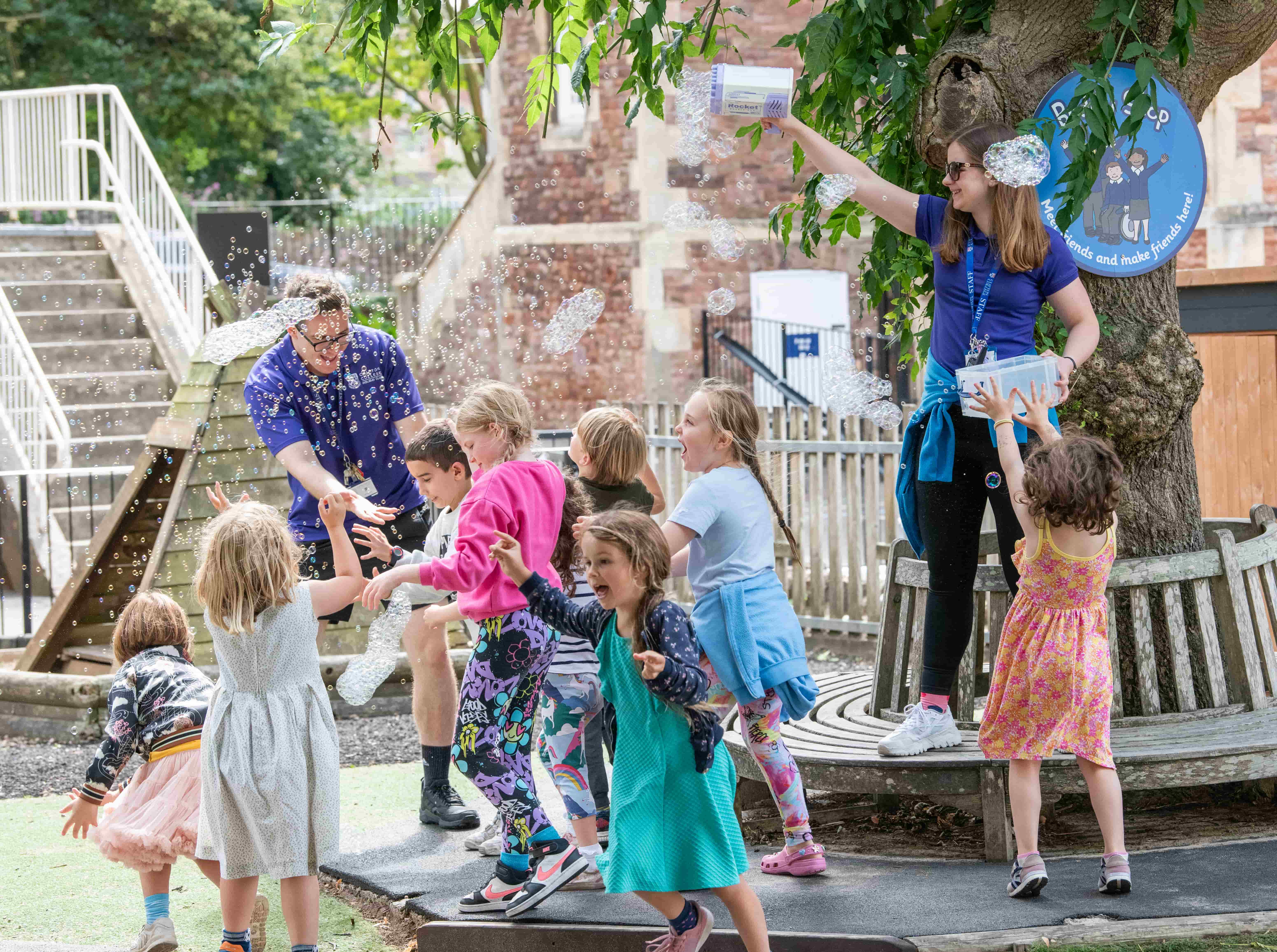 Children joyfully playing with bubbles outdoors, guided by two adults under a large tree. Steps and a brick building are in the background.
