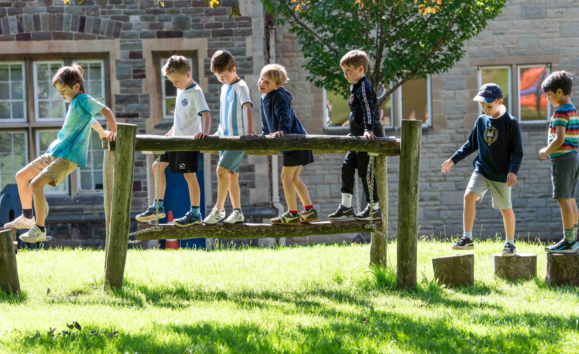 Seven children balance on wooden beams in a sunny park, with a stone building and trees in the background.