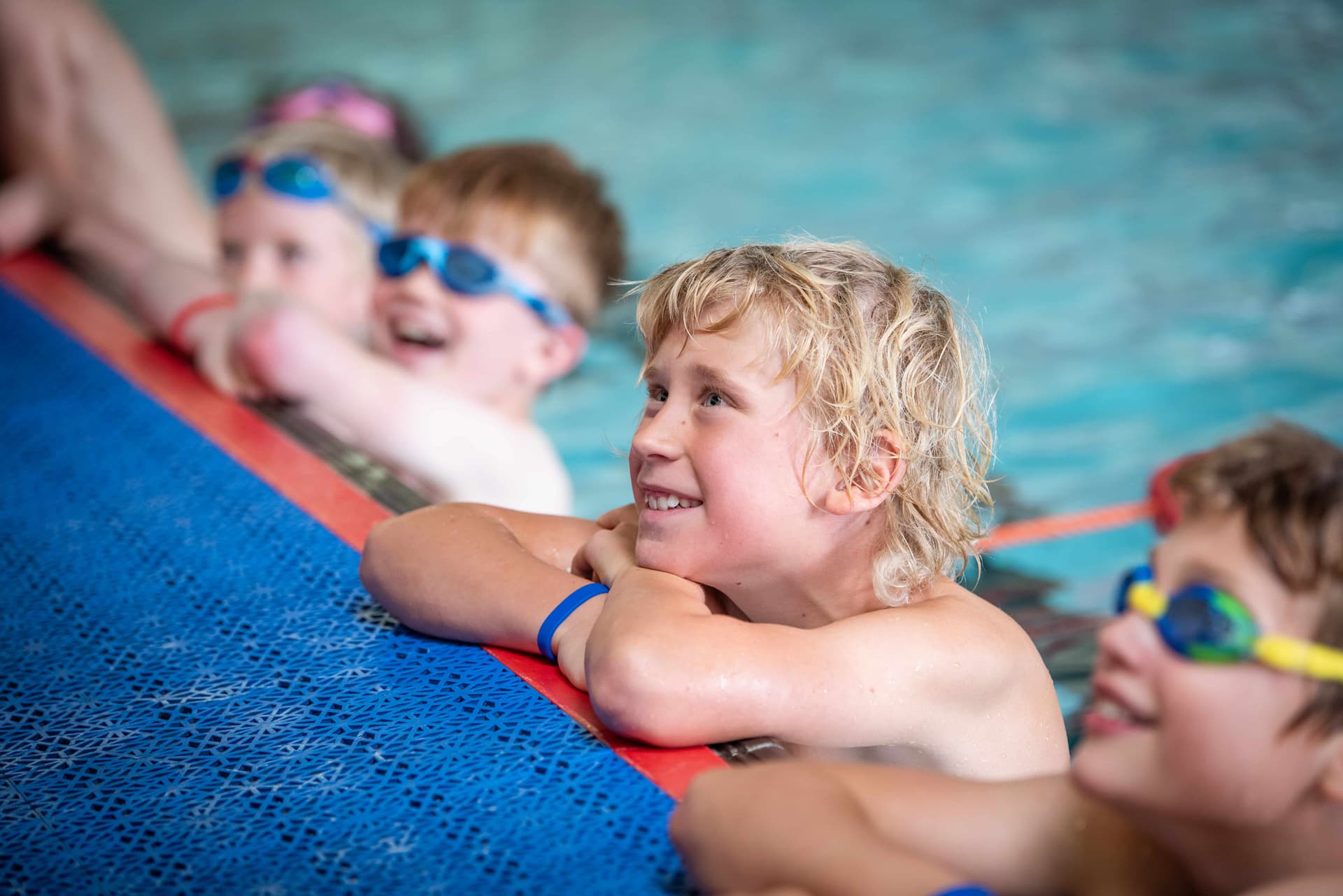 Children in a swimming pool, wearing goggles, smiling and leaning on the pool edge, with water in the background.