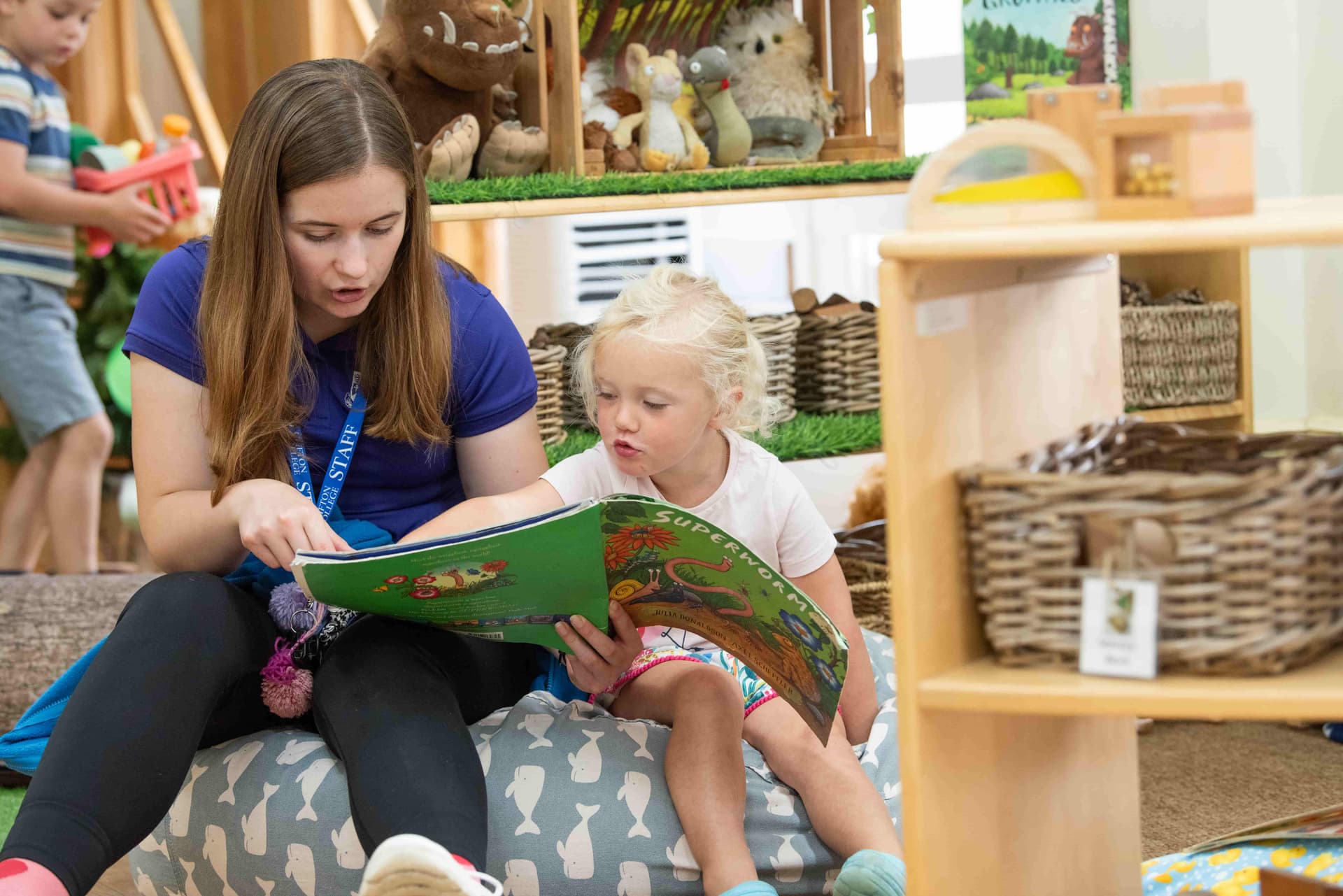 Young woman reading a colorful book to a small child in a cozy, book-filled room with stuffed animals and wooden shelves.