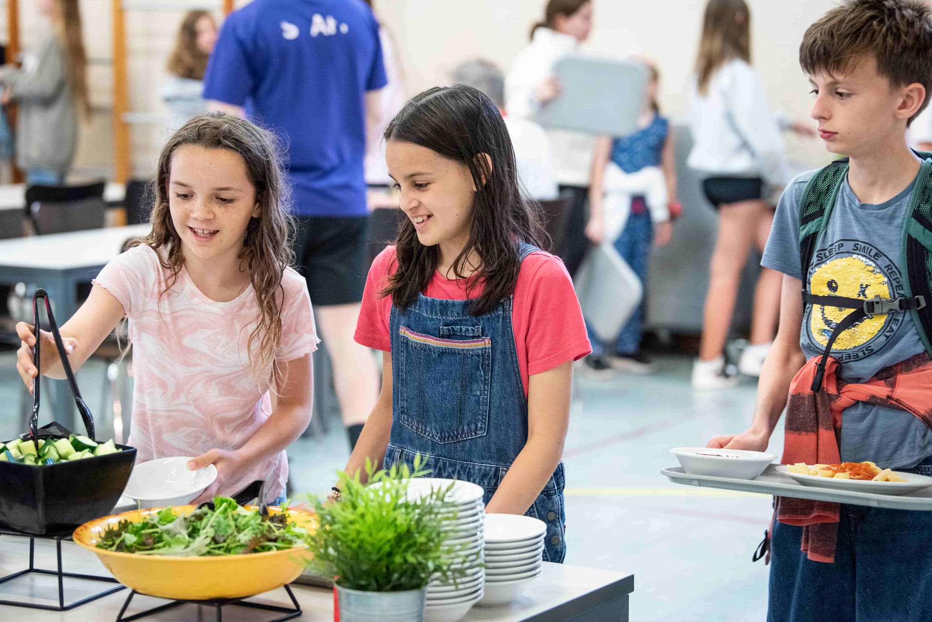 Children in a cafeteria line, serving themselves salad from a buffet. They are smiling and chatting, with others in the background.